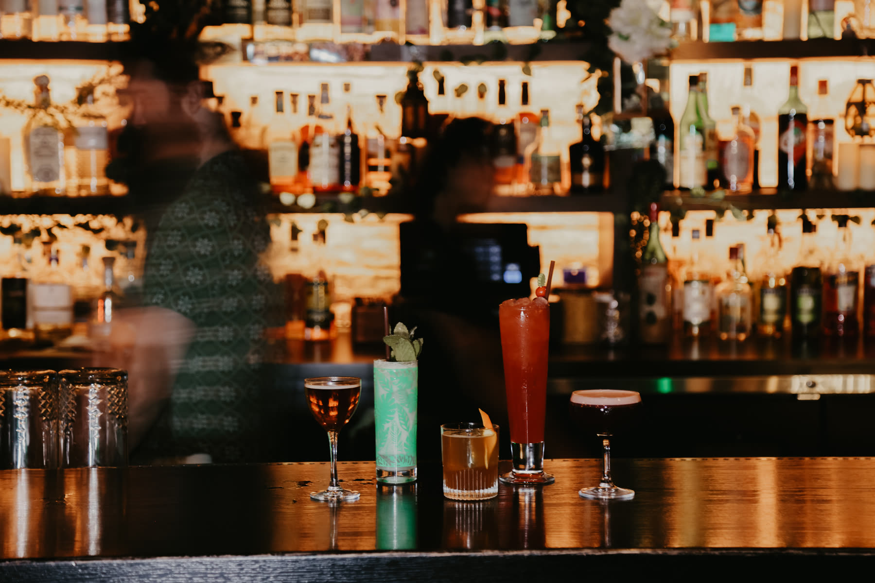 Five cocktails lined up along the bar top with the lit back bar behind